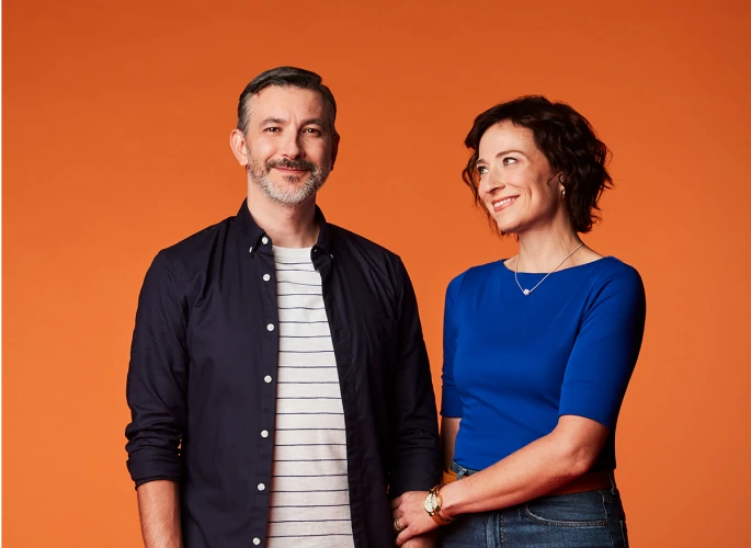 A middle aged couple where the man is smiling at the camera and the woman is smiling supportively at the man