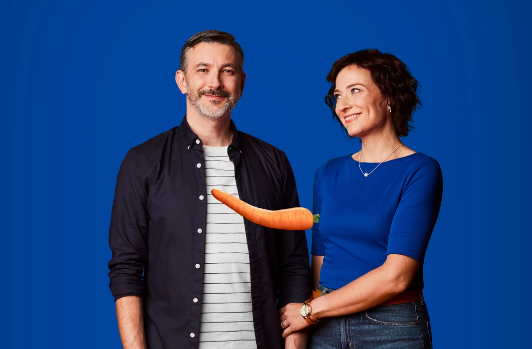 A middle aged couple where the man is smiling at the camera and the woman is smiling supportively at the man – there’s a single bent carrot suspended in the middle of the picture