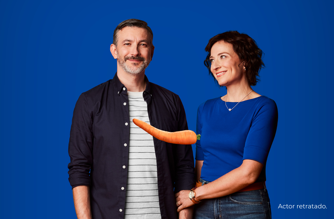 A middle aged couple where the man is smiling at the camera and the woman is smiling supportively at the man – there’s a single bent carrot suspended in the middle of the picture