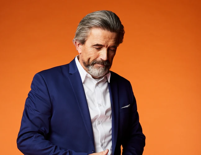 An older man standing against an orange background and looking down